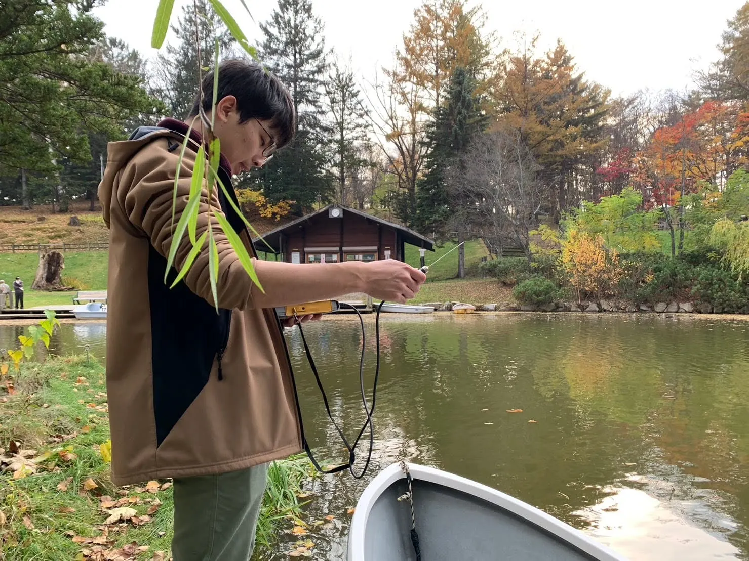 紅葉した木々に囲まれた池のほとりで、ボートのそばに立ち、気温計を手に測定しているところ