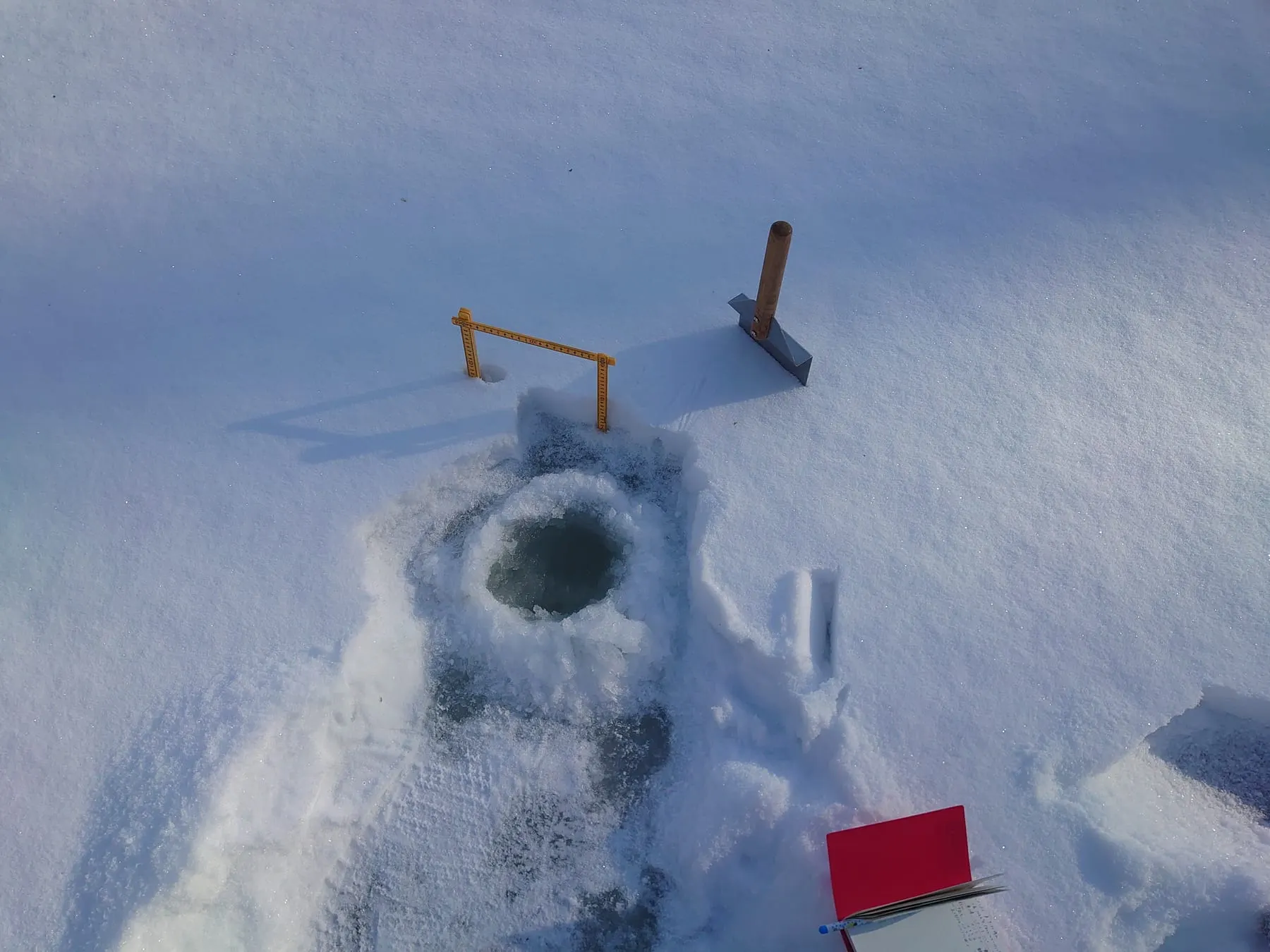 雪に覆われた氷に開けられた丸い穴と、そばに置かれたスコップ、アイスドリル、赤い野帳