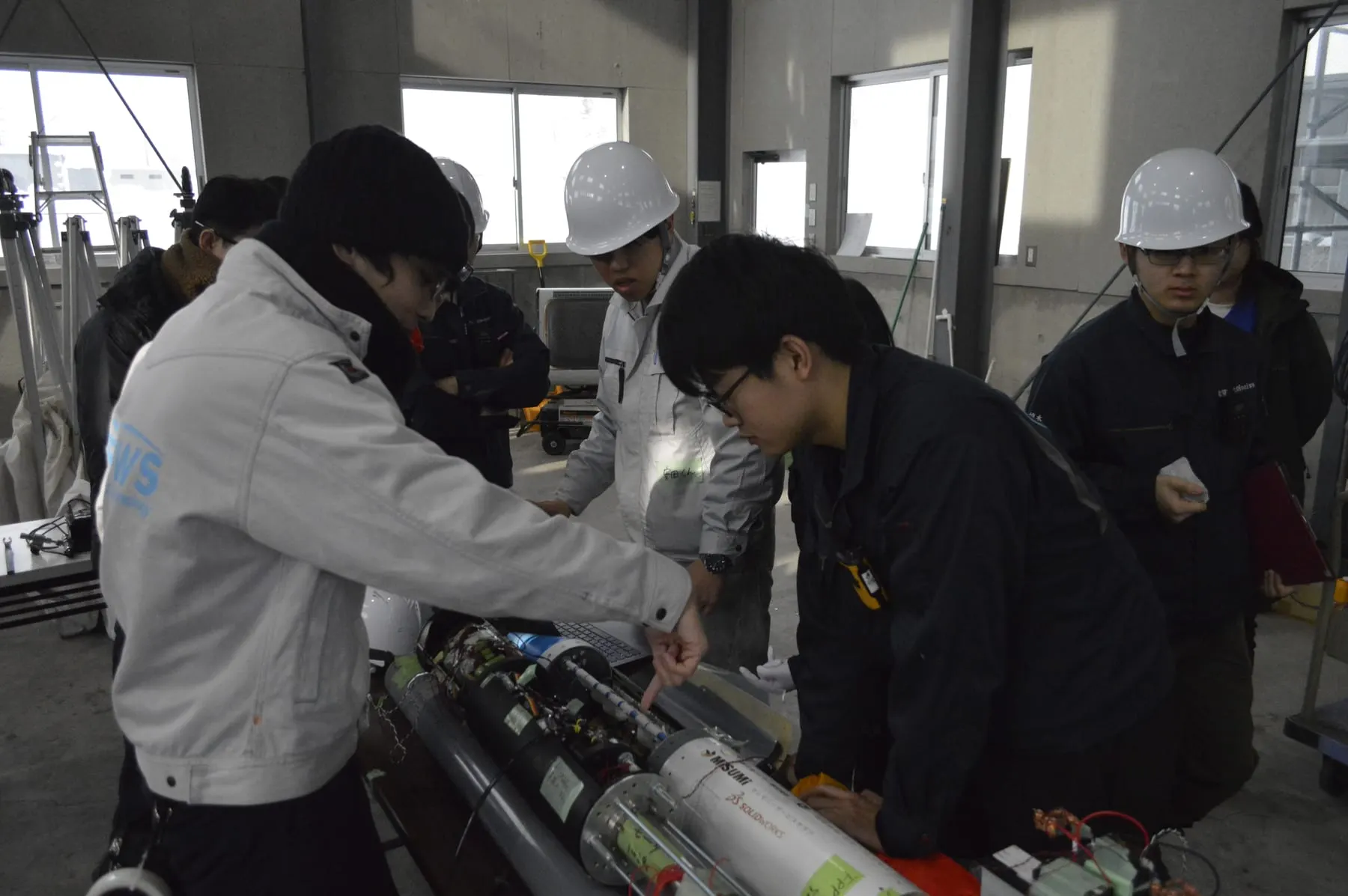 作業台の上のハイブリッドロケットの機体を、ヘルメットをかぶった学生数人が囲んで整備しているところ